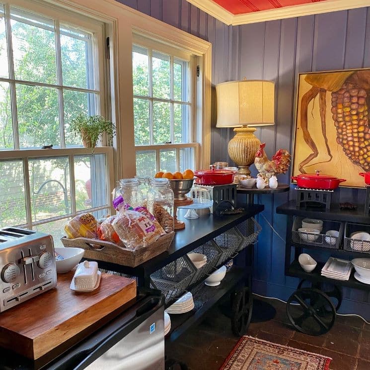 A bright, cozy kitchen featuring a variety of food items, shelves, and a decorative painting on the wall.