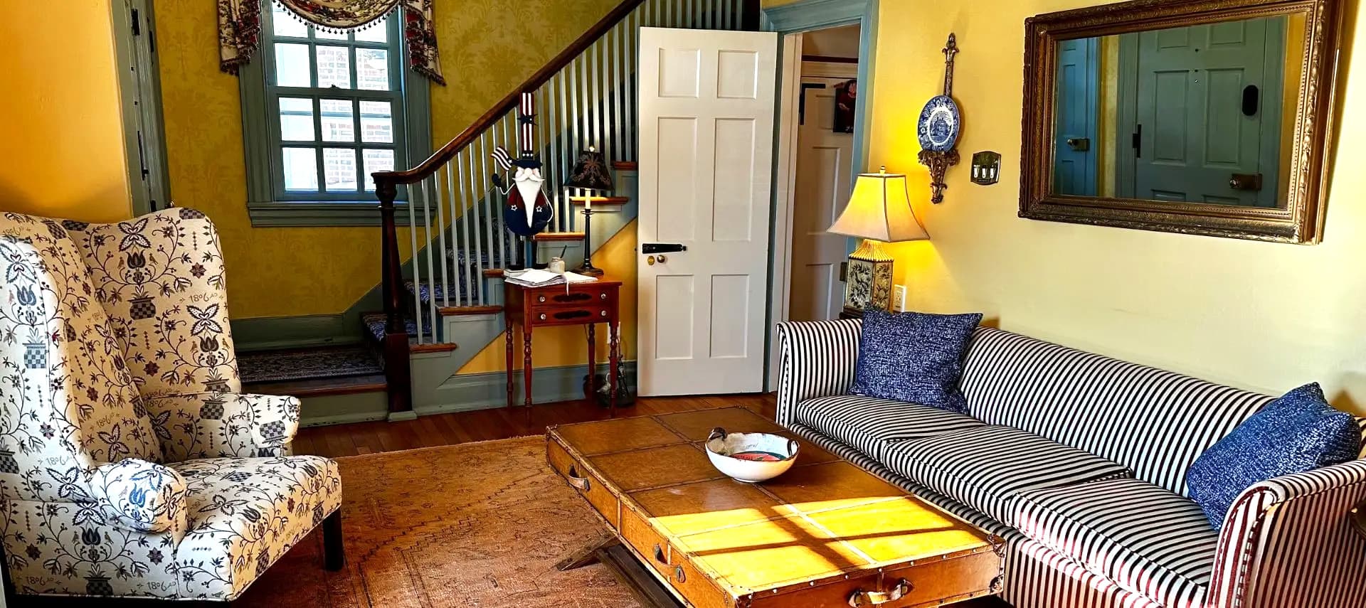 A cozy living room featuring a striped sofa, patterned armchair, and wooden coffee table, with yellow walls and a staircase in the background.