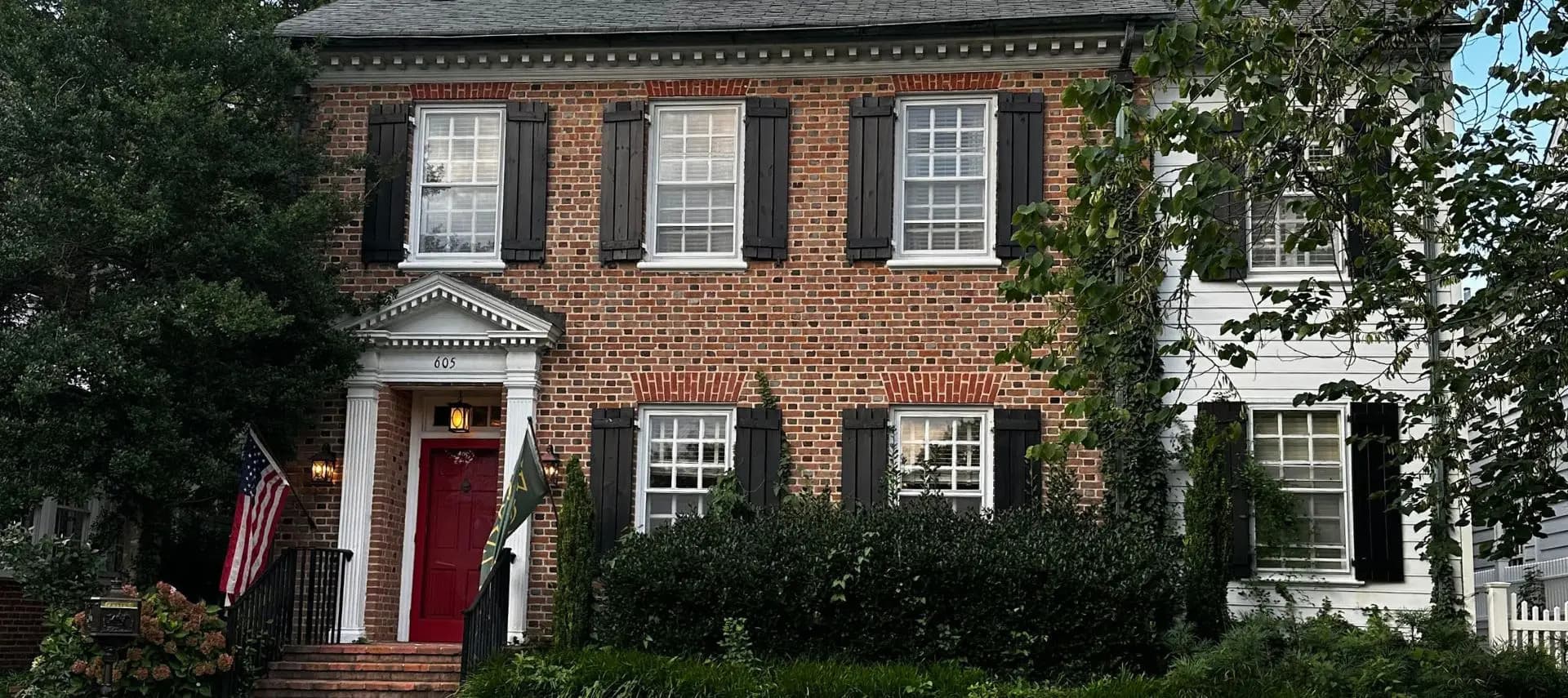 A brick two-story house with white trim, a red door, and an American flag in front.